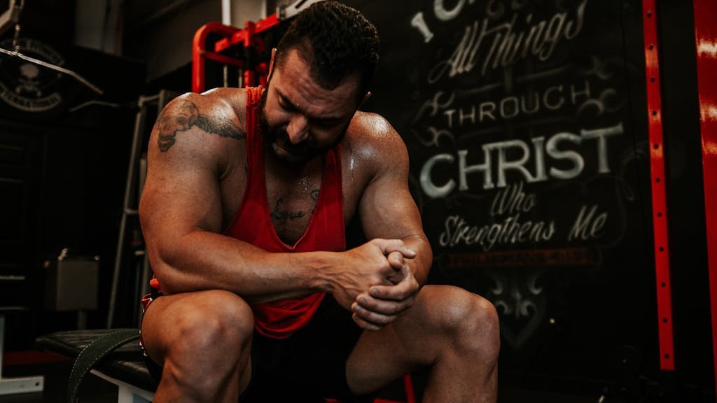 Strong, muscular man in red tank top sitting indoors with inspirational tattoos, showcasing strength and fitness.